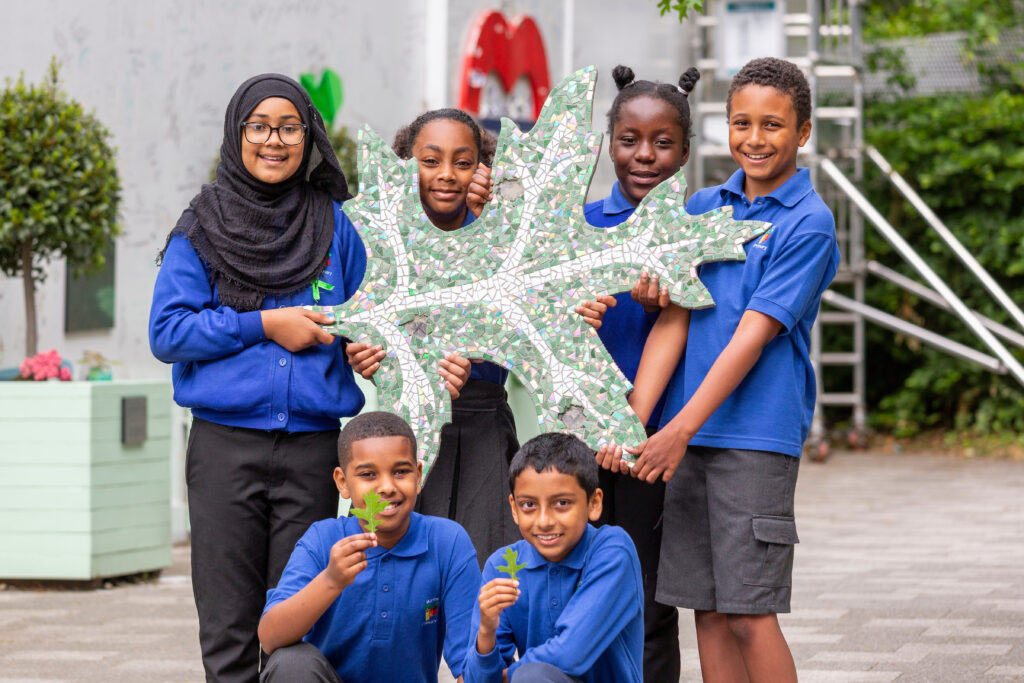 Six children in school uniform pose holding a large mosaic leaf, behind them the hoarding at the base of Grenfell Tower.
