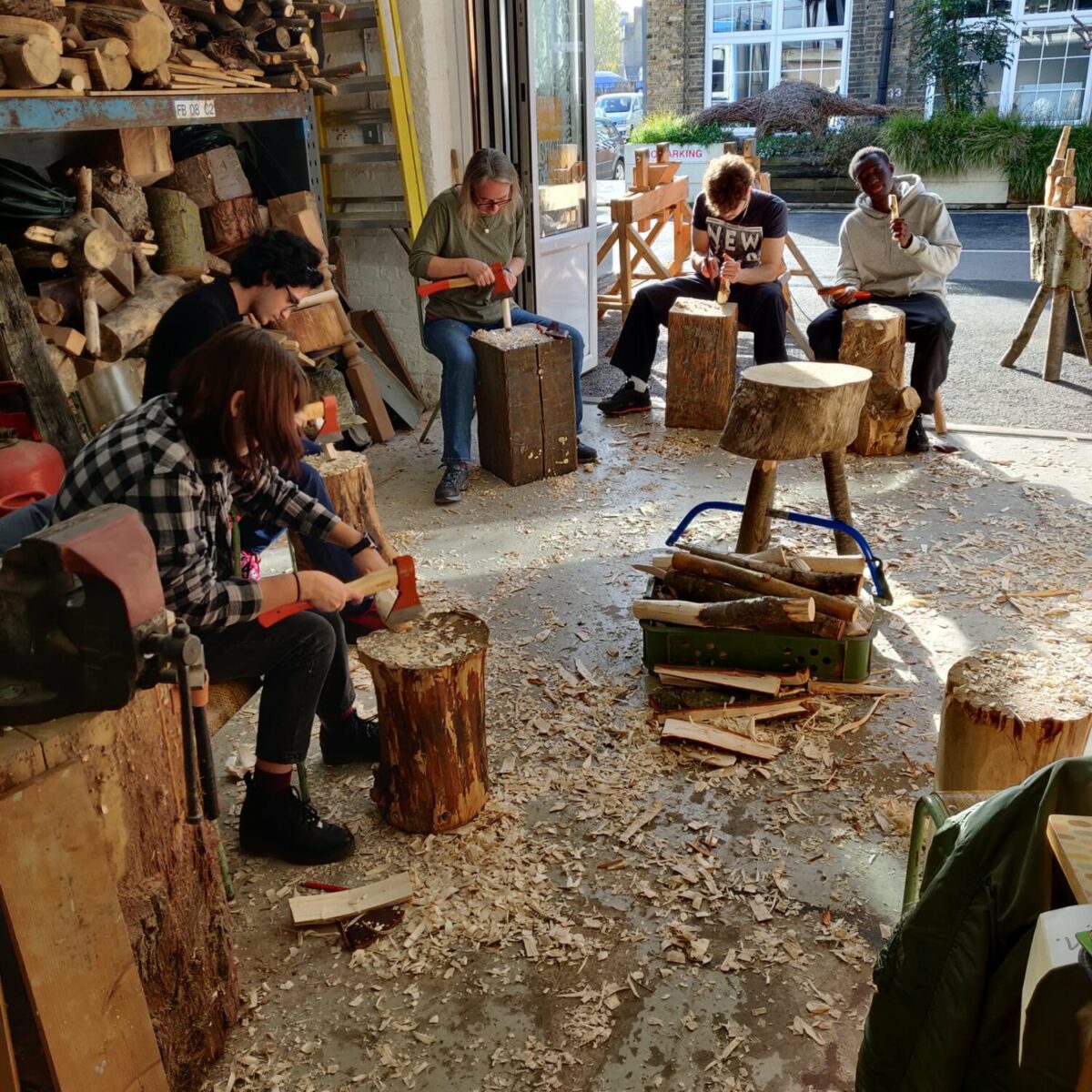 View of a spoon carving workshop, participants sit around a large workshop using axes and chisels on small pieces of wood