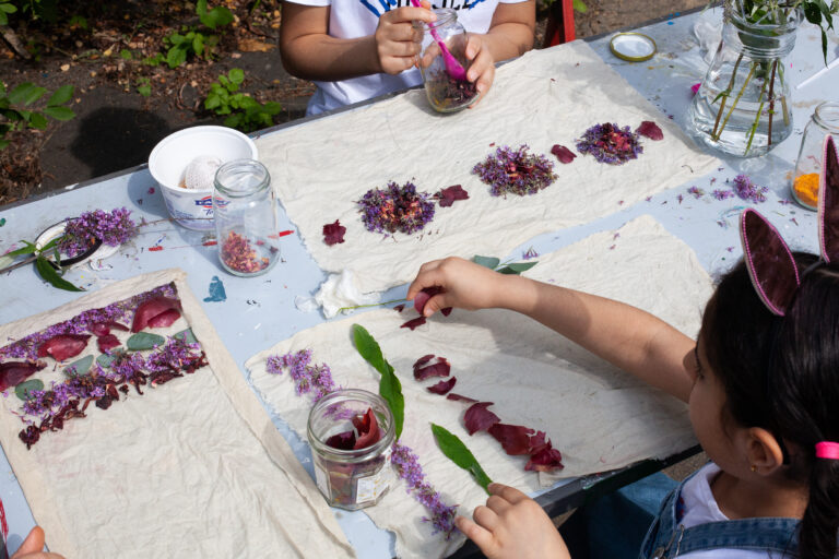 Close up photograph of two children placing dried petals and leaves on fabric during a natural dye workshop, around a table, outdoors.