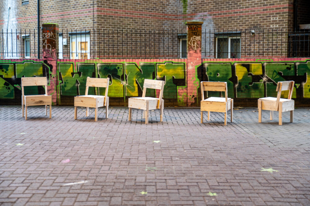 Photograph of five wooden Enzo Mari inspired chairs lined up against a graffiti decorated brick wall, behind a brick building.