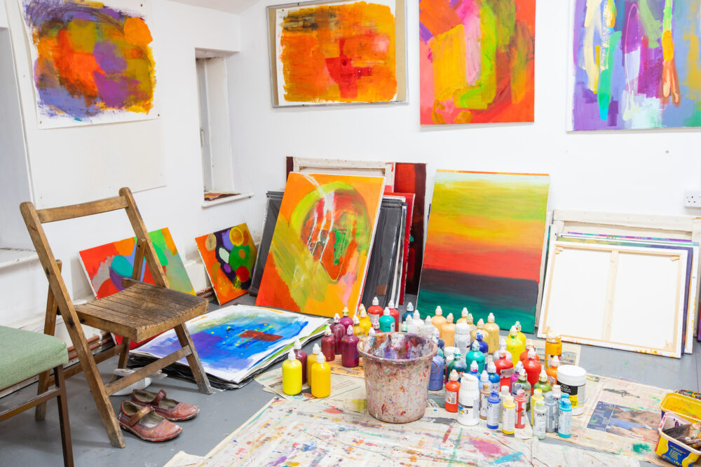 Photograph taken inside a painter's studio, painted canvas stacked against the walls, acrylic paint tubs on the floor.