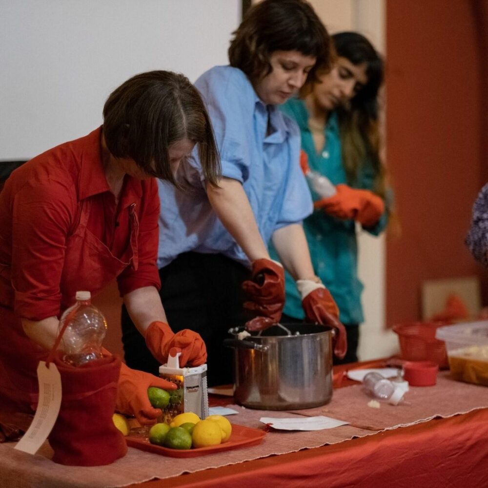 Closeup photograph of two people cooking around a red table, one wearing red and grating lemons and the other wearing blue and using a potato mash, both wearing large red rubber gloves.