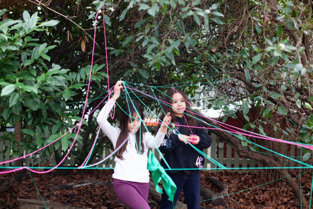 Photograph of two children playing outdoors in a park, weaving colourful string around trees and bushes.