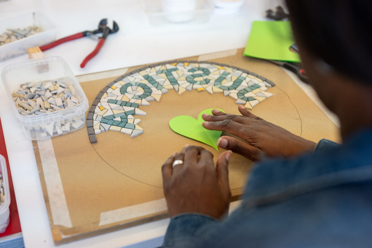 Close-up photograph of a person moving a green paper heart to the centre of a mosaic roundel, whilst they work on the piece, on a workshop table.