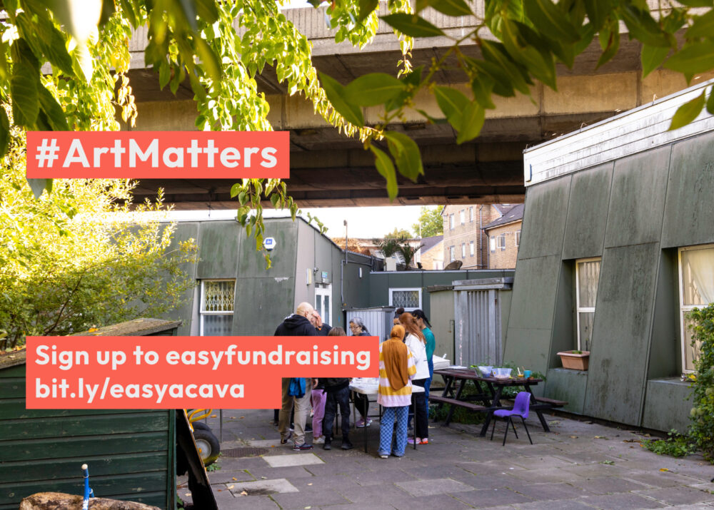 A group of people gather outdoors under the Westway surrounded by greenery. A banner reads #ArtMatters and theres a sign encouraging sign-ups for easyfundraising at the provided link.