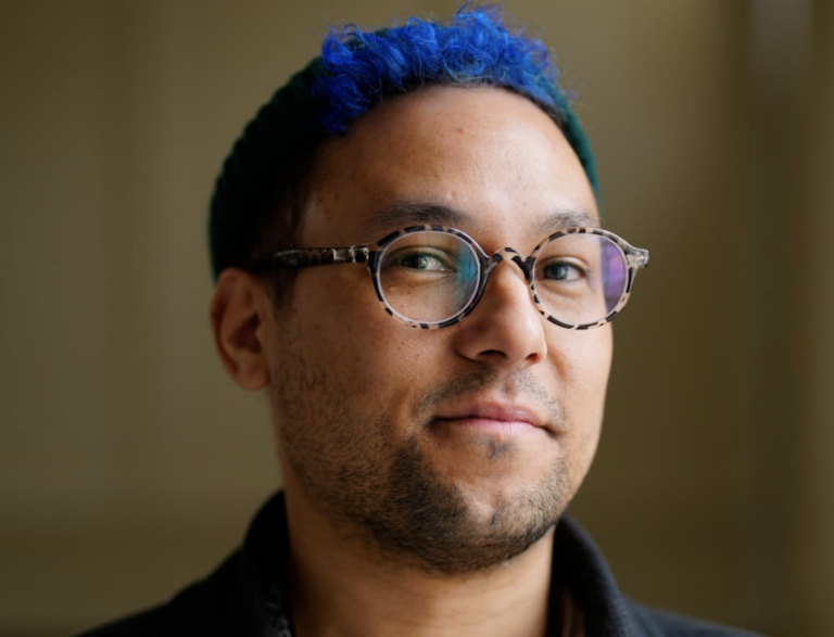 A headshot of Nick with short, curly blue hair, wearing glasses, looking at the camera with a slight smile. They are wearing a dark shirt, and the background is softly blurred.